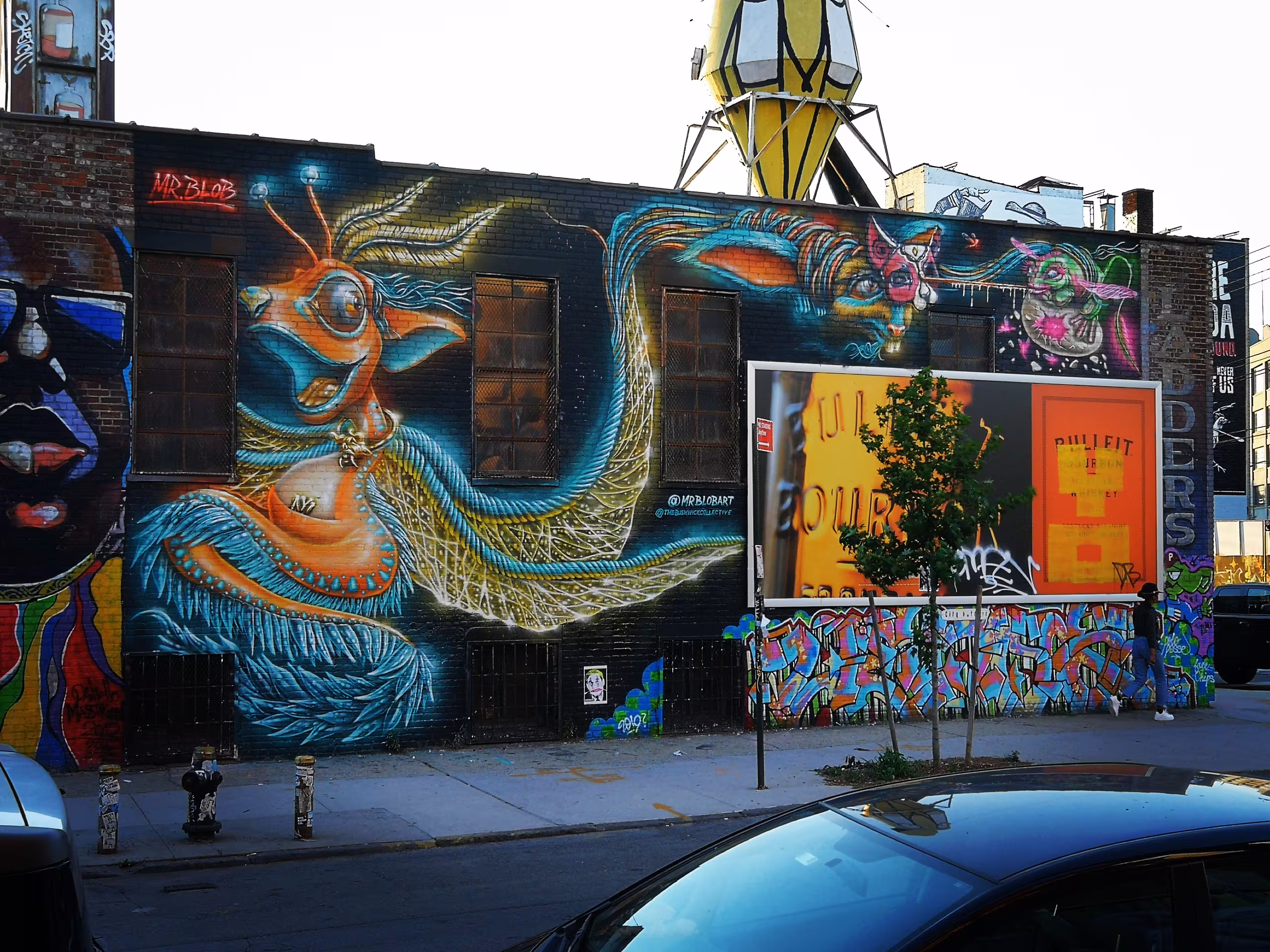 Vibrant horizontal street art mural by Italian artist Mr Blob at The Bushwick Collective in Brooklyn, New York, featuring an orange surreal dragon character stretching across a dark brick factory wall.