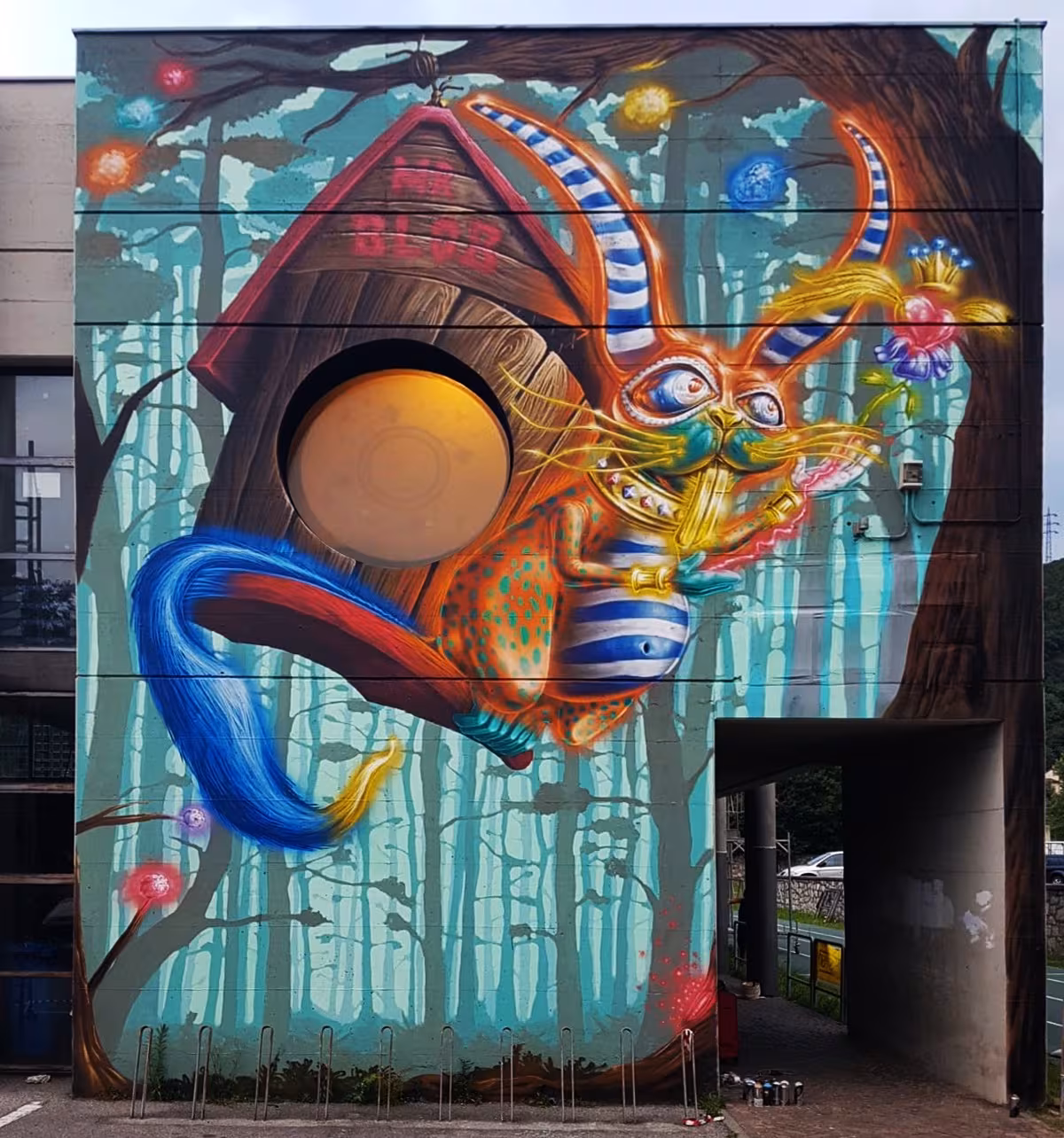 Stunning pop surrealism graffiti mural by Mr Blob integrating an architectural circular window into a wooden birdhouse, featuring a psychedelic cheshire cat character against a misty forest background.